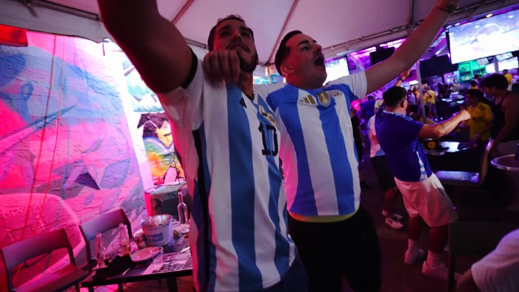 Two Argentina soccer fans in blue and white jerseys cheer with arms raised during a match watch party at Grails Miami.