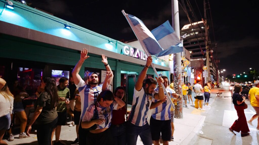 Argentina fans in Argentina jerseys waving the national flag and cheering outside Grails in Wynwood at night