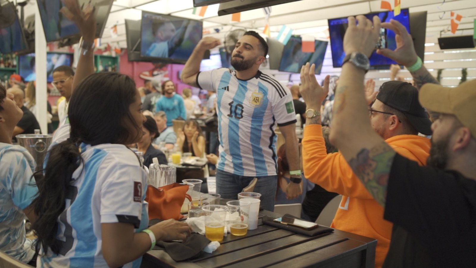 Argentina fan in a Messi #10 jersey celebrates a goal as friends cheer and film at Grails sports bar in Miami.