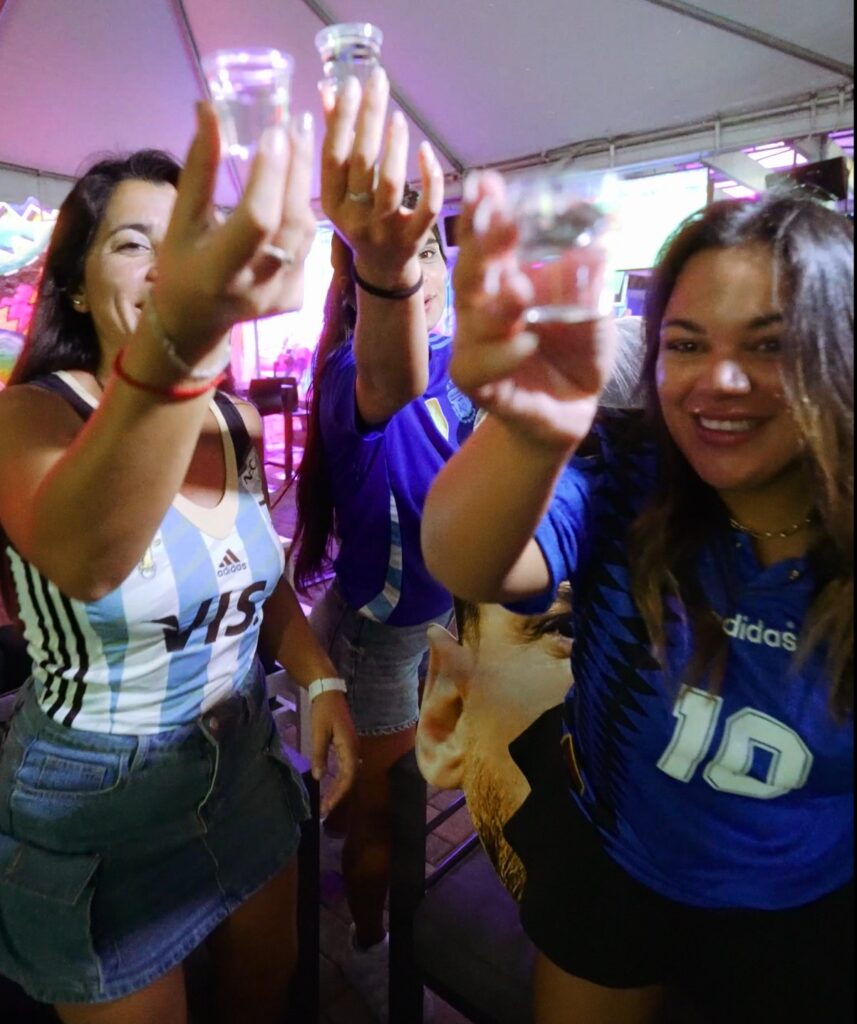 Three women in Argentina soccer jerseys hold up empty shot glasses toward the camera under a tent at Grails Miami.