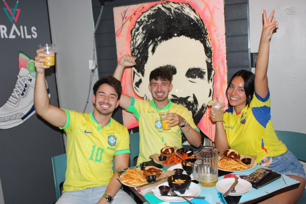 Three friends in Brazil and Ecuador soccer jerseys raise drinks and cheer in front of a Messi mural at Grails Miami