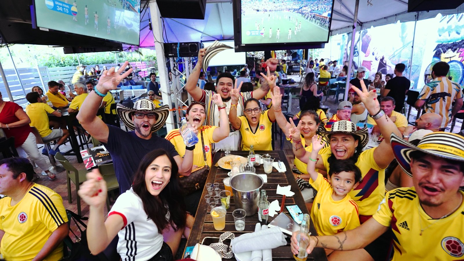 Colombia soccer fans in yellow jerseys and vueltiao hats cheer at a packed table during the Copa America final watch party at Grails Miami.