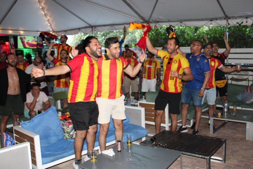Espérance de Tunis soccer fans in red and yellow jerseys cheer and wave scarves during a watch party on the Grails Miami patio.
