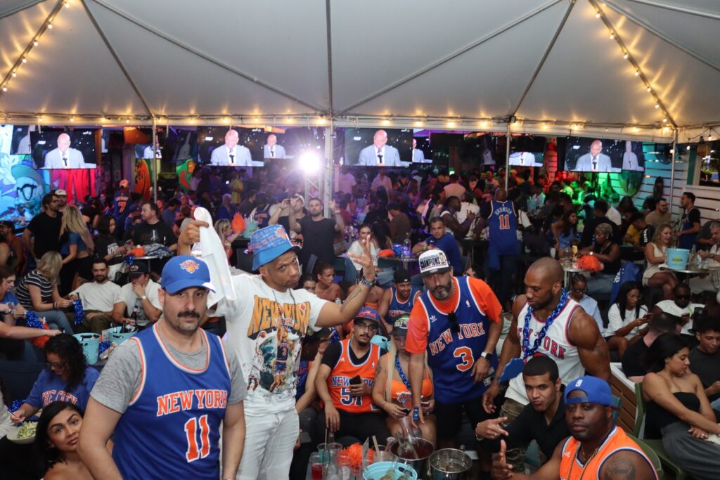 Group of New York Knicks fans in jerseys and team hats packs the tented patio at Grails Miami during an NBA watch party.