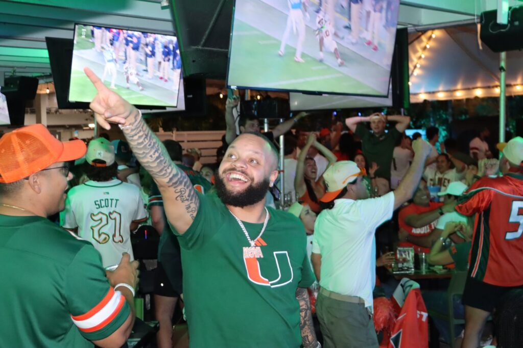 Bearded Miami Hurricanes fan in orange-and-green U shirt points at TV, celebrating during a game watch party at Grails Miami.