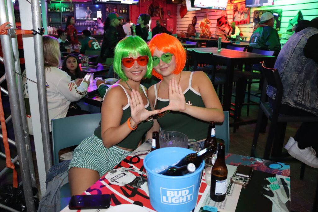 Two young women in green and orange wigs and Miami Hurricanes tank tops throw up 'The U' hand sign at a Grails watch party table with a Bud Light bucket of beers.