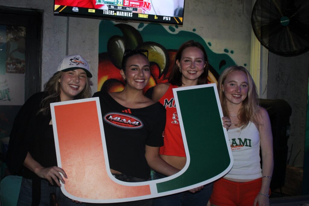 Four smiling young women pose together holding a large Miami Hurricanes U logo sign at a sports bar watch party.