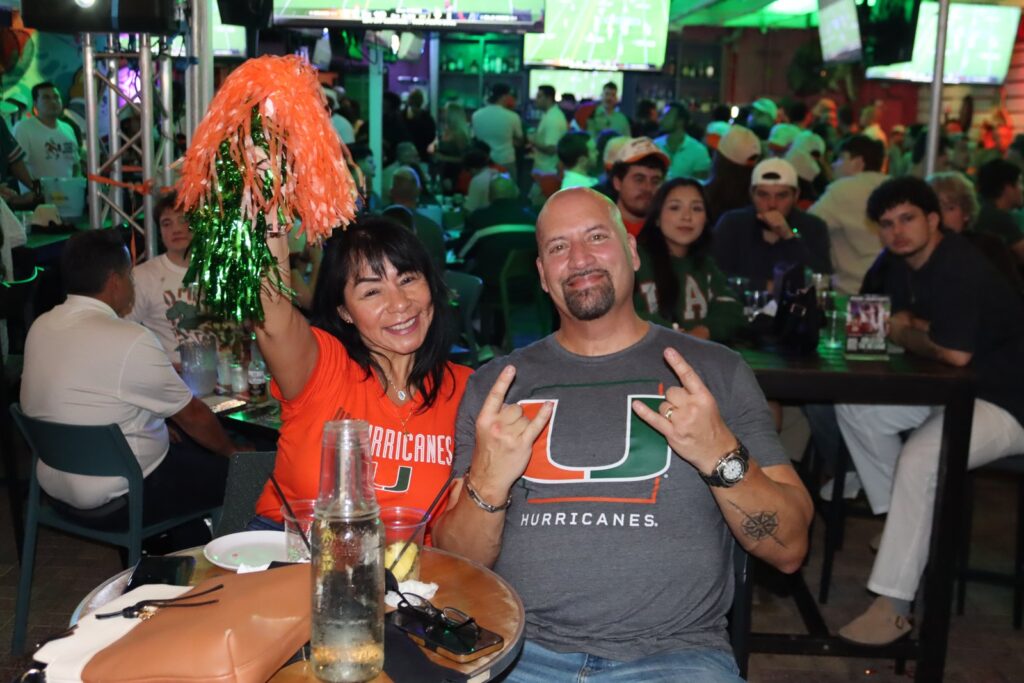 Miami Hurricanes fans in orange and green team gear pose with pom-poms at a packed Grails watch party.