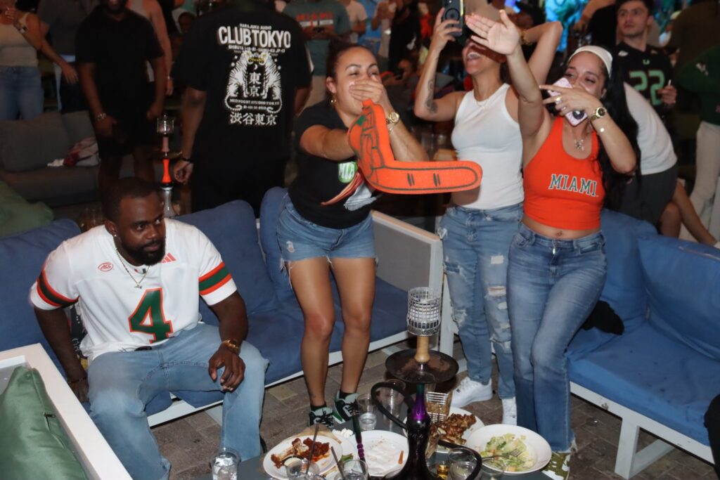 Miami Hurricanes fans in orange and green gear cheer and react during a game watch party at Grails Miami.