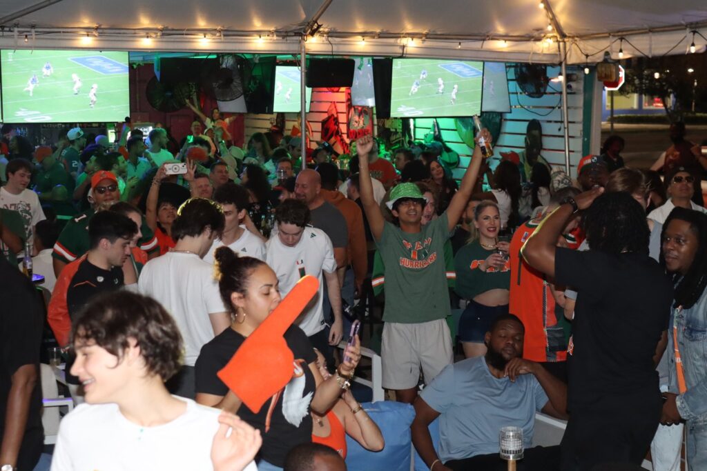 Packed crowd of Miami Hurricanes fans in green and orange jerseys cheering during a football watch party under the Grails tent