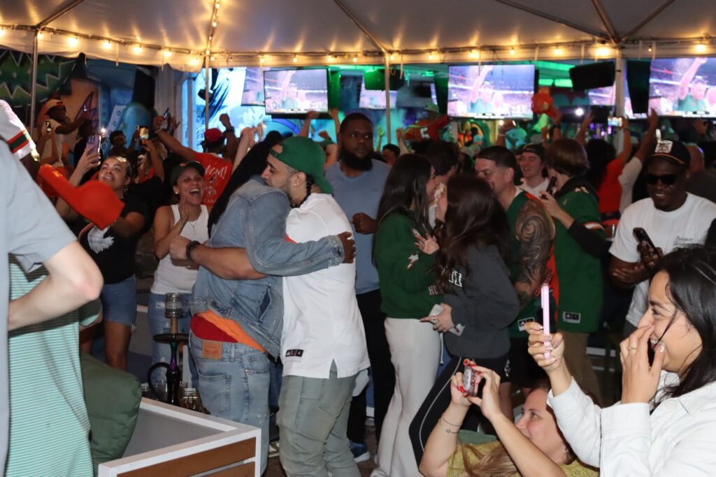 Miami Hurricanes fans in orange and green gear hug and cheer during a football watch party under the tent at Grails Miami