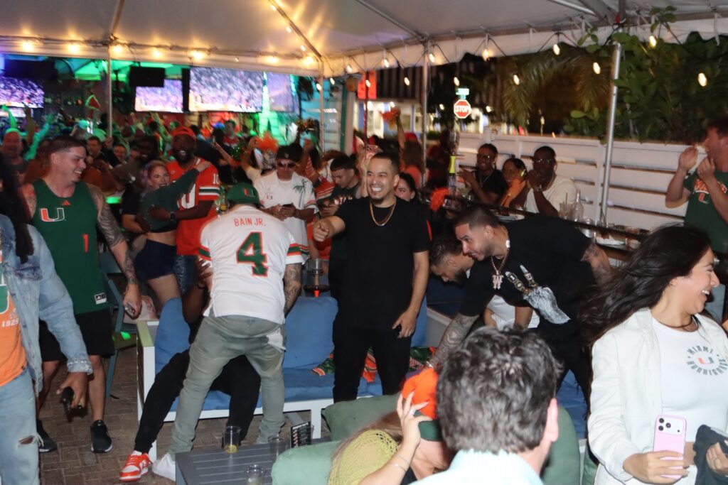 Miami Hurricanes fans in orange and green jerseys cheer and celebrate during a football watch party on the covered patio at Grails Wynwood.